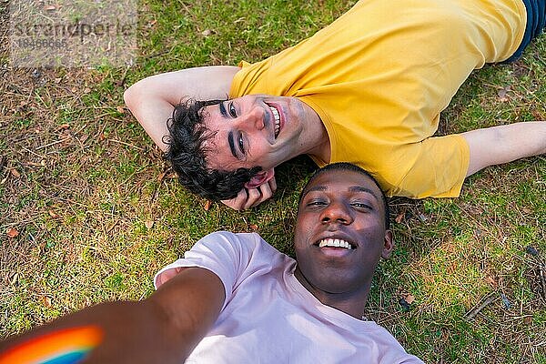 Porträt eines Paares multiethnischer Männer in einem Park  Igbt Konzept  die auf dem Gras liegen und ein Selfie machen