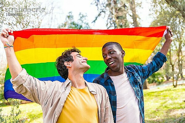 Multiethnisches schwules männliches Paar  das eine Regenbogenflagge mit dem Symbol der Homosexualität hält und Spaß hat