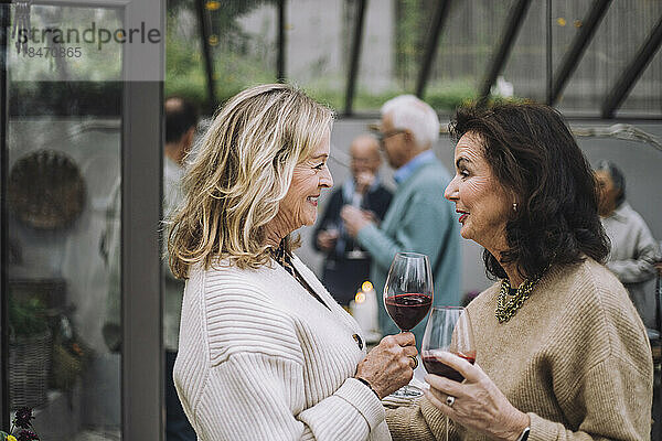 Lächelnde ältere Frau mit Weingläsern im Gespräch mit Freundinnen bei einer Dinnerparty