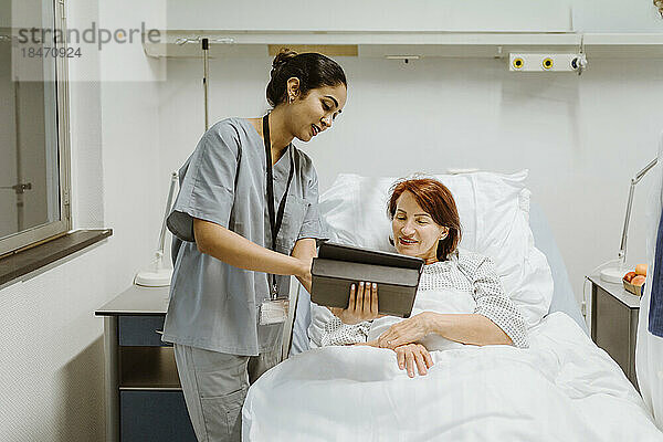 Eine lächelnde Krankenschwester zeigt einer älteren Patientin  die auf einem Bett im Krankenhaus liegt  ein digitales Tablet