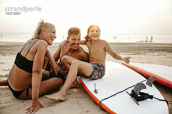 Fröhliche Eltern genießen mit Tochter sitzen auf Paddleboard am Strand bei Sonnenuntergang