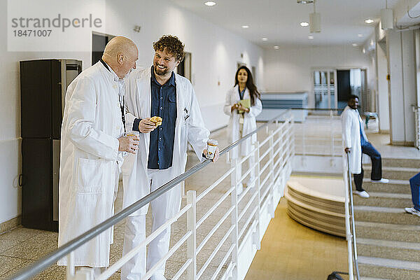 Fröhliche männliche Ärzte  die sich während der Kaffeepause im Krankenhausflur am Geländer stehend unterhalten