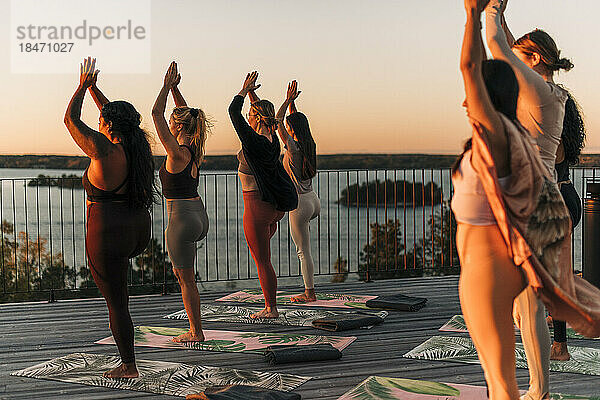 Weibliche Freunde üben zusammen auf der Terrasse bei Sonnenuntergang eine Baumpose