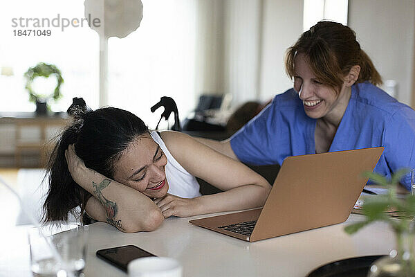 Glückliche Betreuerin im Gespräch mit einer querschnittsgelähmten Frau  die zu Hause auf ihren Laptop schaut