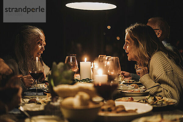 Ältere Frau im Gespräch mit einer Freundin am Esstisch während eines Abendessens bei Kerzenschein