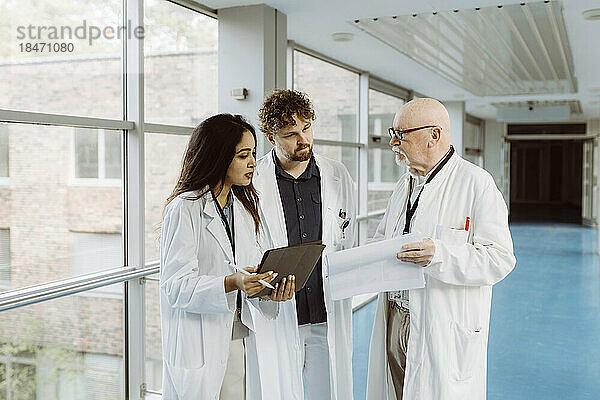 Kollegen aus dem Gesundheitswesen diskutieren über ein digitales Tablet und ein Dokument  während sie im Krankenhausflur zusammenstehen