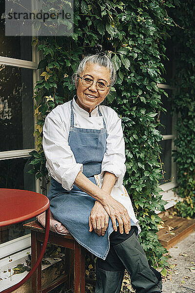 Porträt einer lächelnden Senior-Köchin  die vor einem Restaurant sitzt