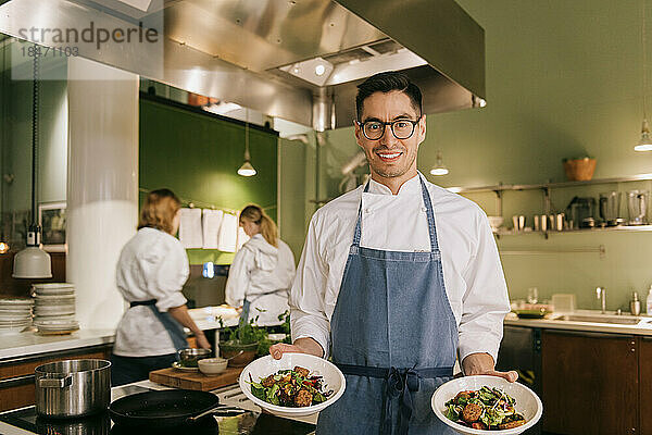 Lächelnder männlicher Koch  der Schüsseln mit Lebensmitteln hält  während er in einer Restaurantküche steht