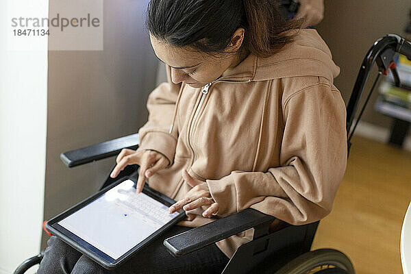 Hochformatige Ansicht einer querschnittsgelähmten Frau  die zu Hause im Rollstuhl sitzend einen Tablet-PC benutzt