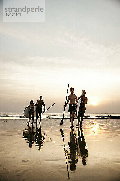 Familie mit Paddelbrettern zu Fuß gegen den Himmel bei Sonnenuntergang am Strand