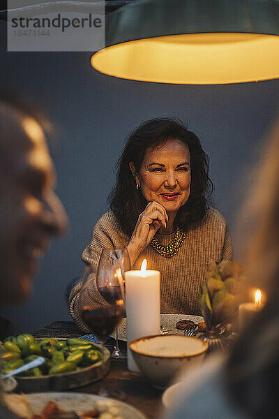 Lächelnde ältere Frau mit Hand am Kinn bei beleuchteter Dinnerparty