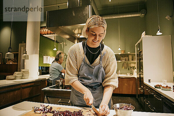 Lächelnde nicht-binäre Person  die während eines Kochkurses in der Küche Gemüse hackt