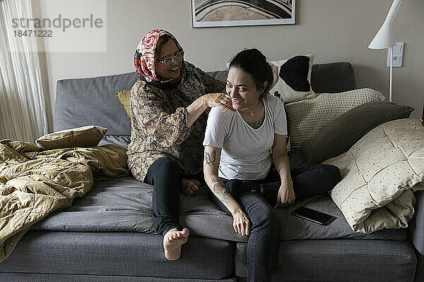 Glückliche Frau mit Querschnittslähmung wird von ihrer Mutter massiert  während sie zu Hause auf dem Sofa sitzt