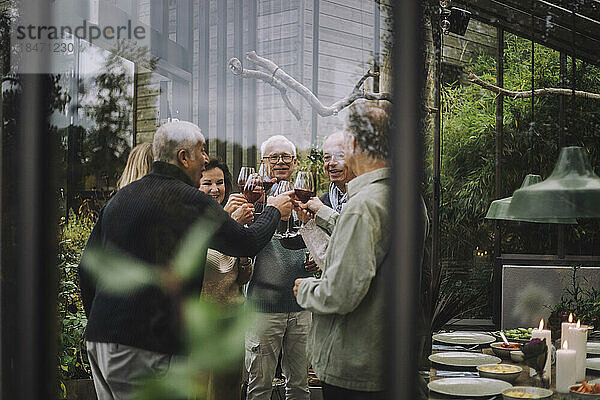 Männliche und weibliche Freunde im Ruhestand stoßen mit Getränken auf einer Party an  gesehen durch Glas