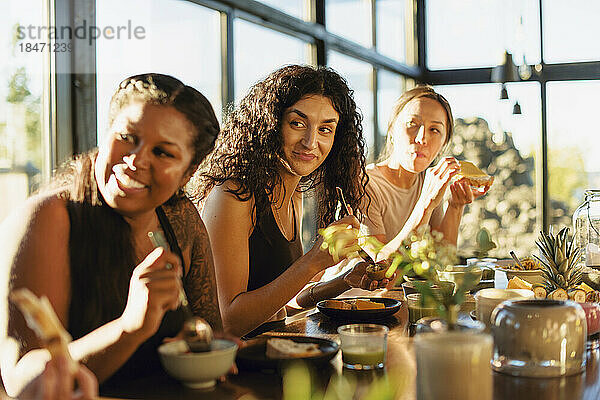 Lächelnde Freundinnen schauen weg  während sie beim Frühstück am Esstisch sitzen