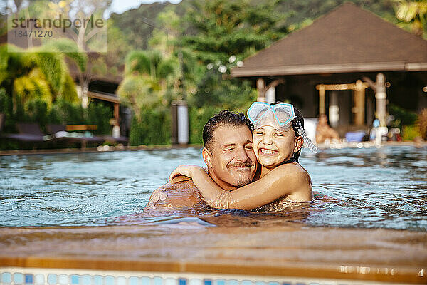 Glückliches Mädchen umarmt Vater beim Schwimmen im Pool am Urlaubsort
