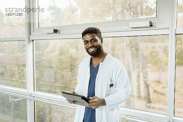 Porträt eines lächelnden jungen Arztes  der ein digitales Tablet hält und am Fenster eines Krankenhauskorridors steht