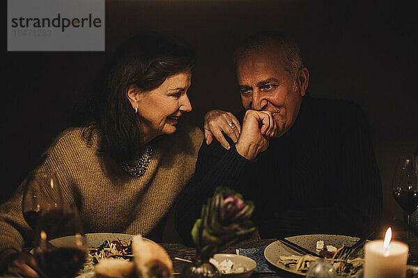 Lächelnde ältere Frau im Gespräch mit reifem Mann mit Hand am Kinn auf einer Dinnerparty