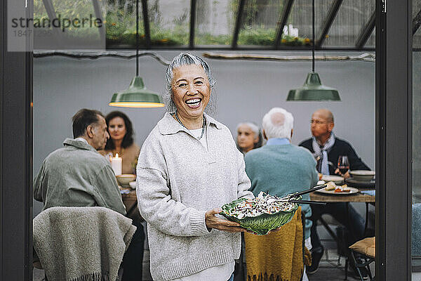 Porträt einer fröhlichen älteren Frau mit einer Salatschüssel in der Hand bei einer Dinnerparty
