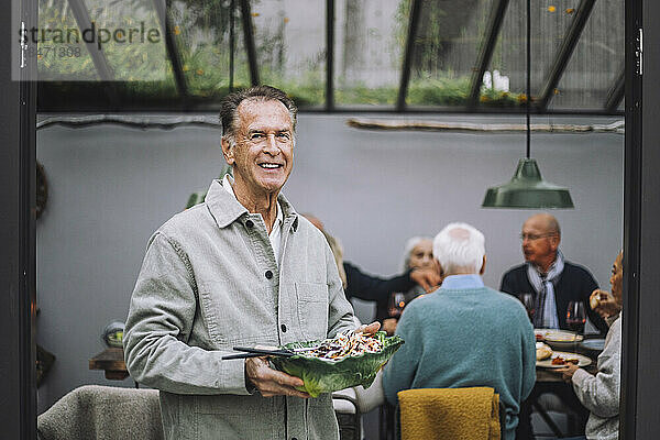 Porträt eines lächelnden älteren Mannes  der eine Salatschüssel bei einer Dinnerparty hält