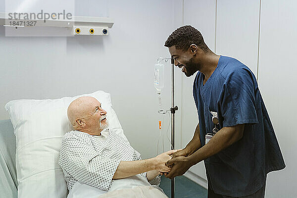 Lächelndes Pflegepersonal hält die Hände eines älteren männlichen Patienten  der auf einem Bett im Krankenhaus liegt
