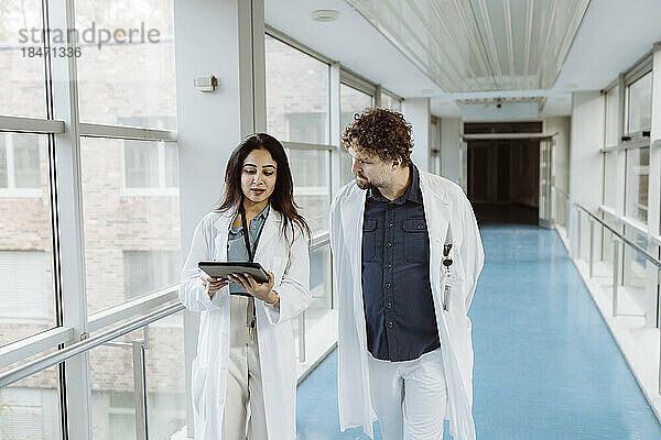 Eine Ärztin diskutiert mit einem männlichen Kollegen über ein digitales Tablet  während sie gemeinsam durch einen Krankenhausflur gehen