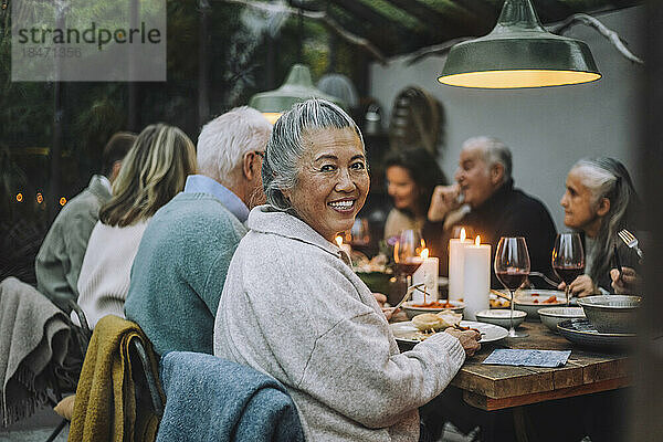 Lächelnde ältere Frau schaut über die Schulter  während sie mit Freunden bei einer Dinnerparty sitzt