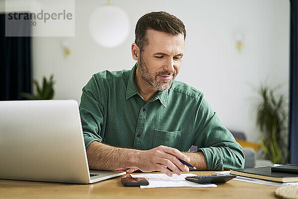 Mann sitzt zu Hause mit Laptop und Dokumenten und prüft die Finanzen seines Hauses