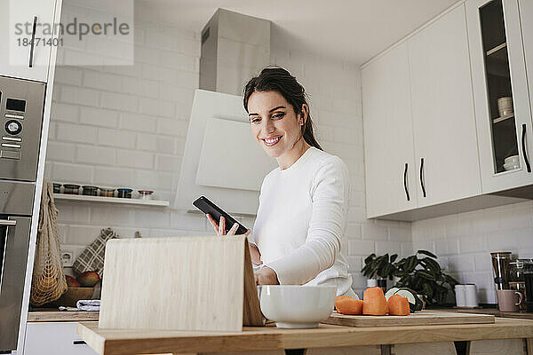 Glückliche Frau benutzt Tablet-PC und hält Telefon in der Küche