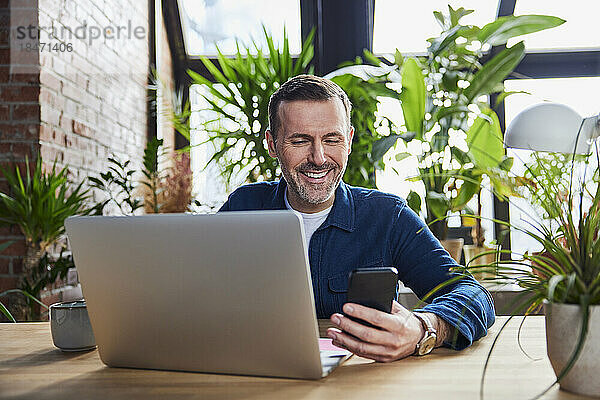 Glücklicher Geschäftsmann mit Laptop und Smartphone im Loft-Büro
