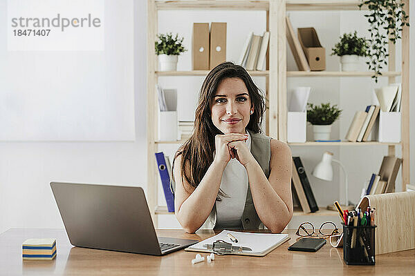 Lächelnde Geschäftsfrau sitzt mit den Händen am Kinn am Schreibtisch im Büro