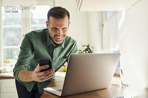 Lächelnder Mann  der in der Küche mit Mobiltelefon und Laptop online bezahlt