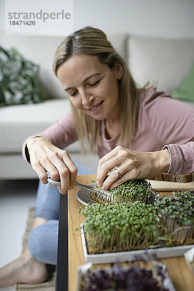 Lächelnde Frau kümmert sich im Wohnzimmer um selbst angebaute Pflanzen