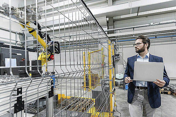 Geschäftsmann mit Laptop steht neben Roboterarm in der Fabrik