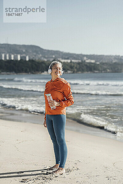 Lächelnde reife Frau mit Wasserflasche steht am Ufer am Strand