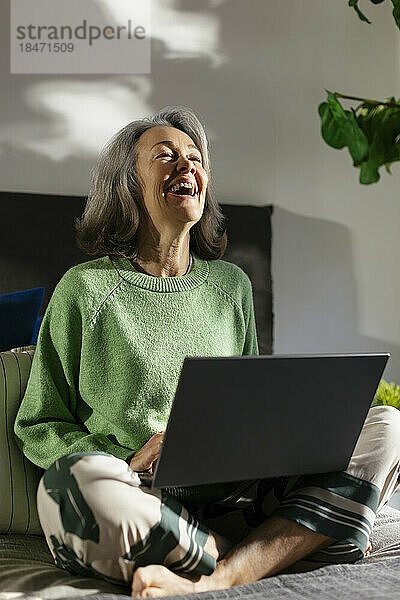 Fröhliche Frau sitzt mit Laptop auf dem Bett im Schlafzimmer