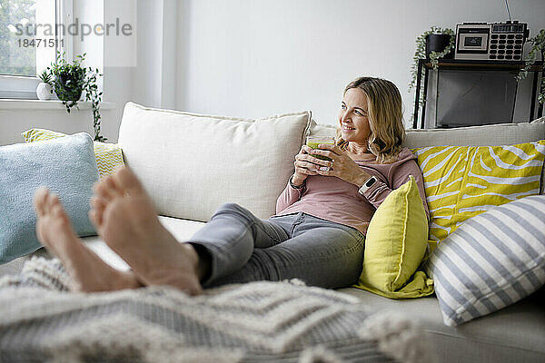 Lächelnde Frau sitzt mit einem Glas Smoothie zu Hause auf dem Sofa