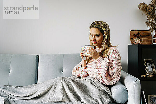 Junge Frau sitzt zu Hause auf dem Sofa und trinkt Kaffee