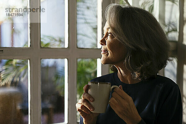 Lächelnde reife Frau mit Kaffeetasse  die durch das Fenster schaut