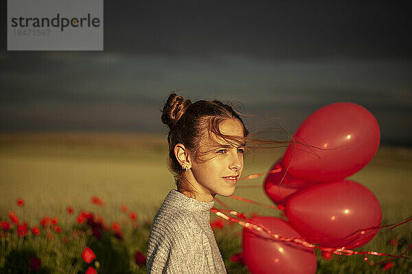 Nachdenklich lächelndes Mädchen steht mit Luftballons am Mohnfeld