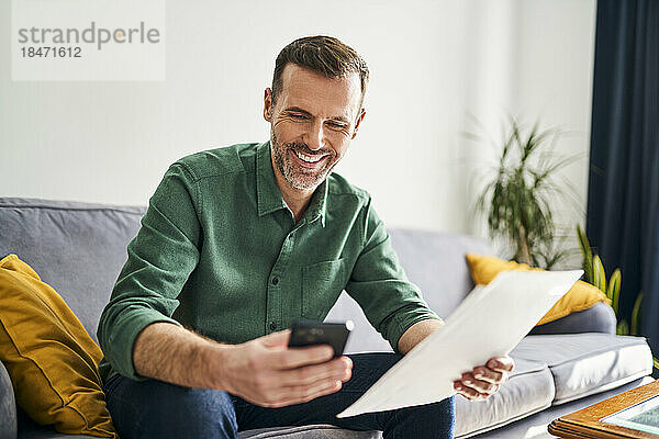 Lächelnder Mann sitzt auf der Couch und bezahlt Rechnungen mit seinem Smartphone