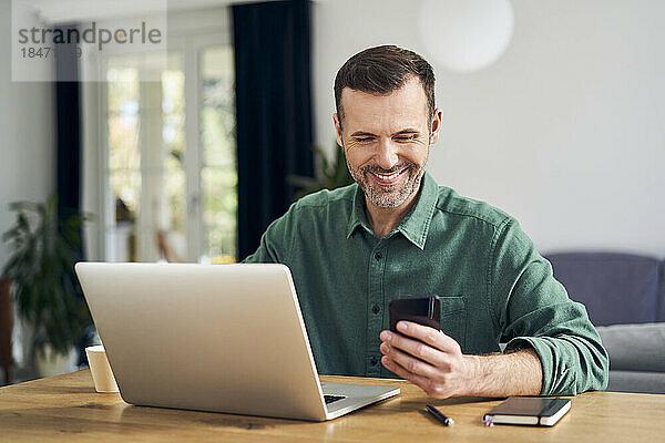 Fröhlicher Mann  der zu Hause am Tisch sitzt und mit dem Smartphone am Laptop bezahlt