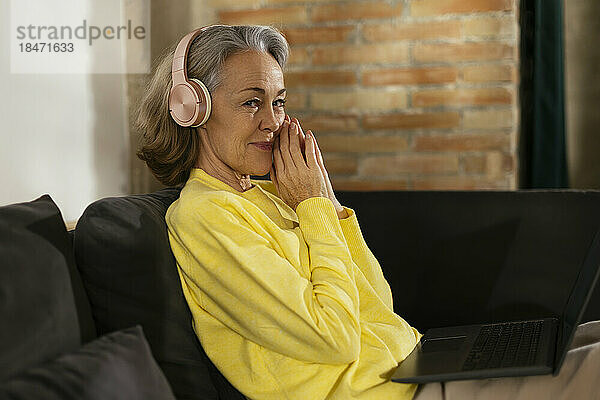 Lächelnder Freiberufler mit kabellosen Kopfhörern sitzt zu Hause auf dem Sofa