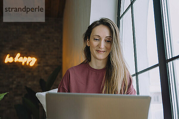 Lächelnde Frau  die zu Hause am Fenster einen Laptop beobachtet