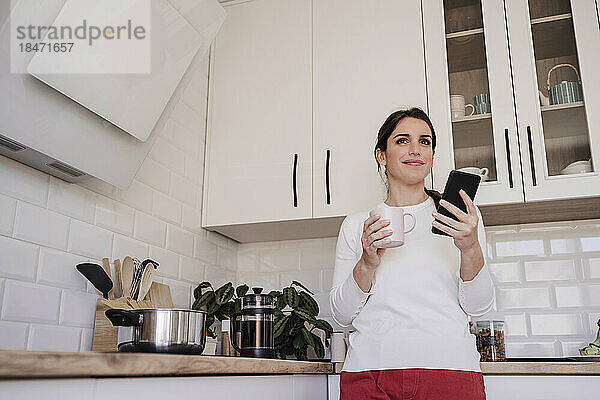Nachdenklich lächelnde Frau mit Smartphone und Kaffeetasse in der Küche