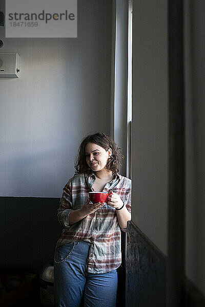 Lächelnde Geschäftsfrau mit Kaffeetasse steht am Fenster im Büro