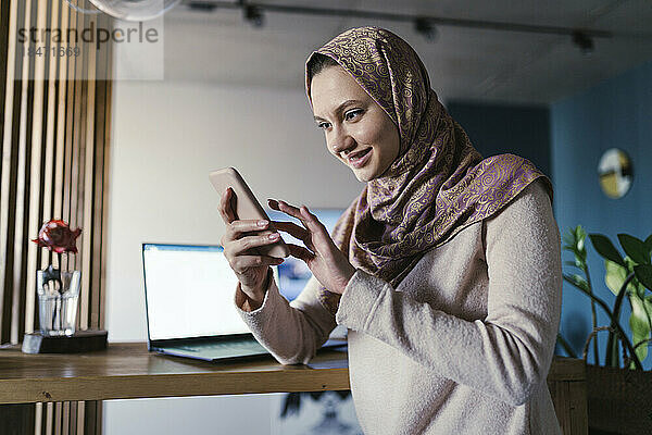 Lächelnder Freiberufler mit Hijab und Smartphone im Heimbüro