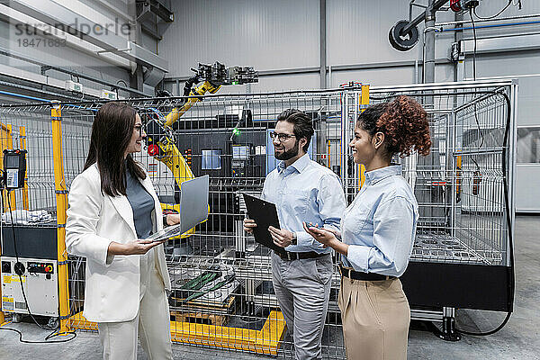 Lächelnde Geschäftsfrau hält Laptop in der Hand und diskutiert mit Kollegen in der Fabrik