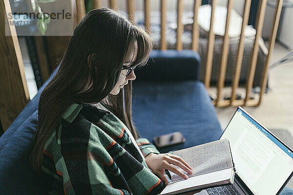 Junge Frau mit Laptop liest zu Hause ein Buch