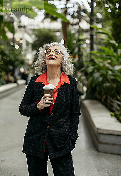 Nachdenkliche reife Geschäftsfrau mit Einwegkaffeetasse steht im Garten